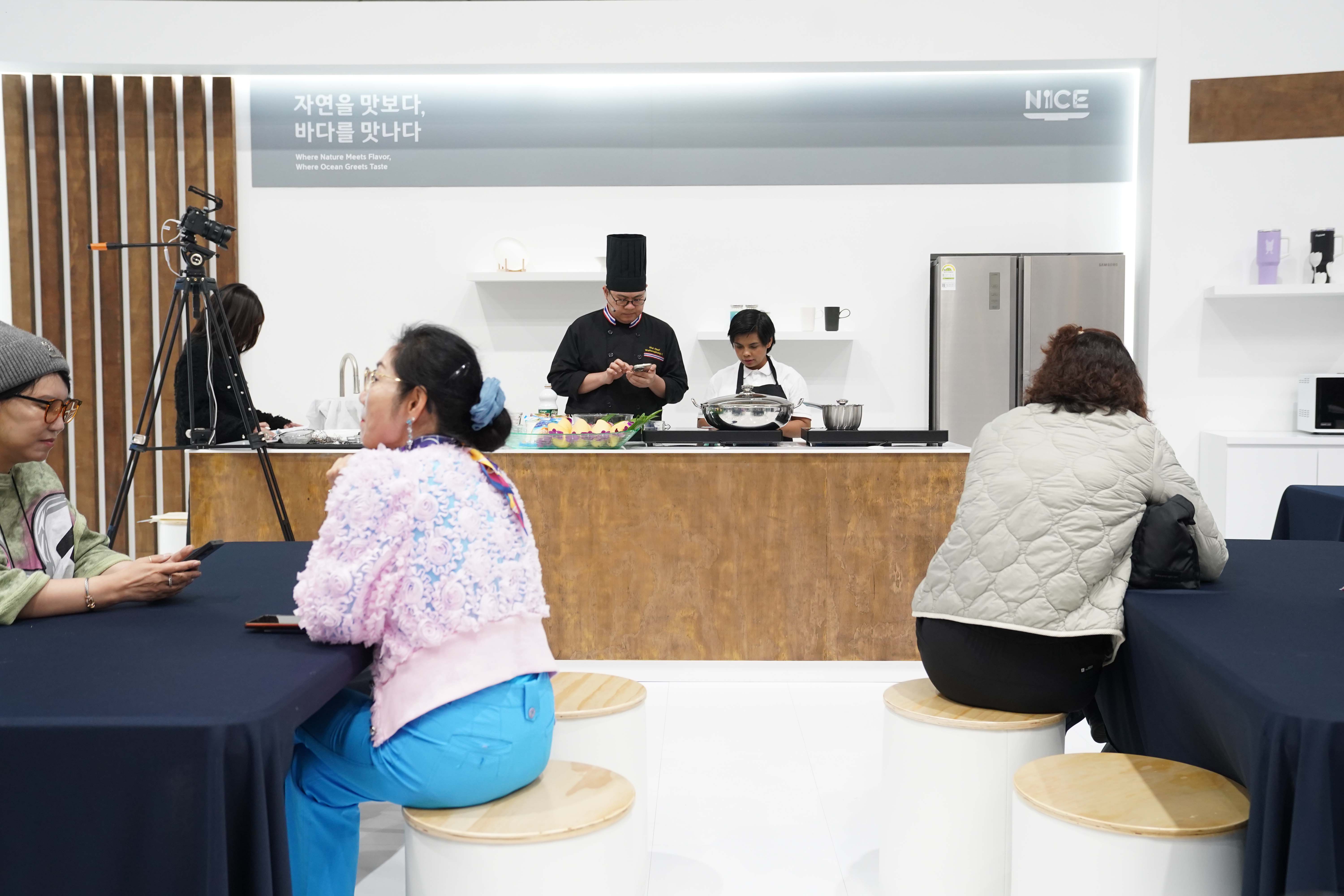 '자연을 맛보다, 바다를 맛보다' 문구와 'NICE' 로고가 적힌 오픈 주방 세트. 셰프들이 요리를 준비하고 있고 앞쪽 테이블에 관람객들이 앉아 있음.