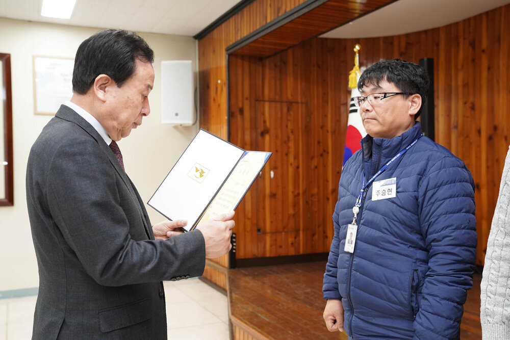 3월 정례조회에서 정장입은 남성이 남색 경량 패딩을 입은 남성에게 패를 건내고 있다.
