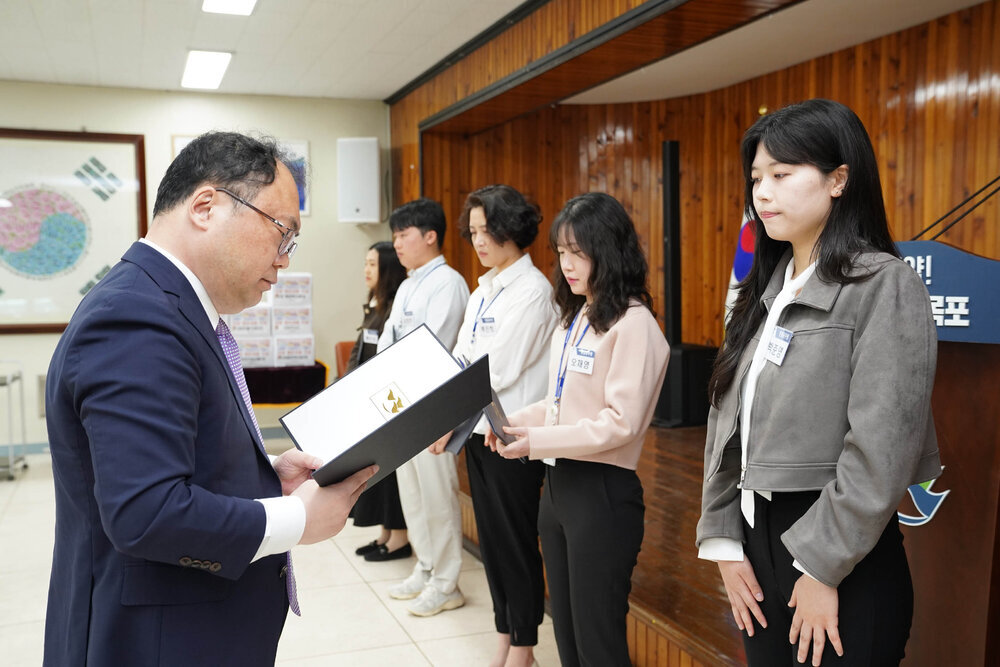 정장을 입고 안경을 낀 남성이 회색 무광 자켓을 입은 여성에게 상장을 수여하고 있다