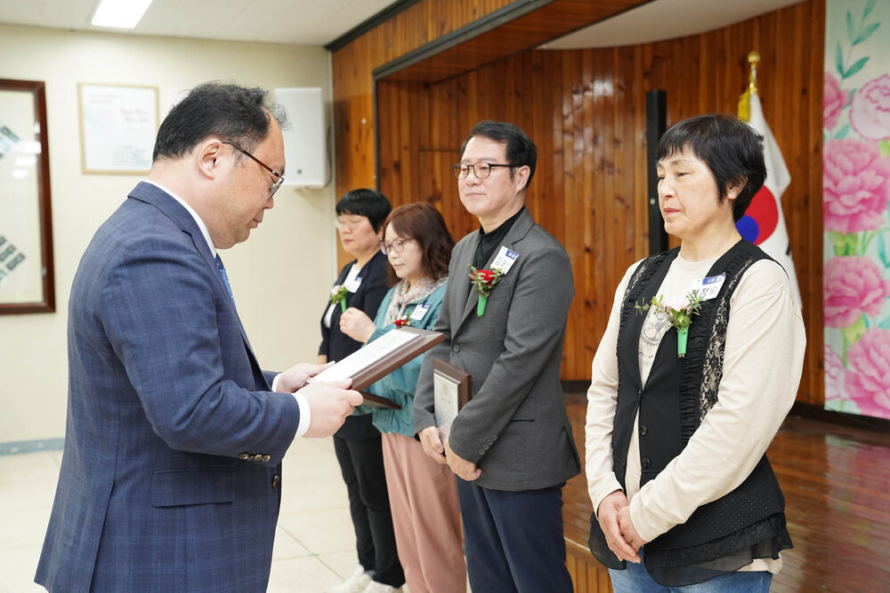 정장을 입고 안경을 낀 남성이 베이지 티에 검은색 베스트를 입은 여성에게 패를 수여하고 있다