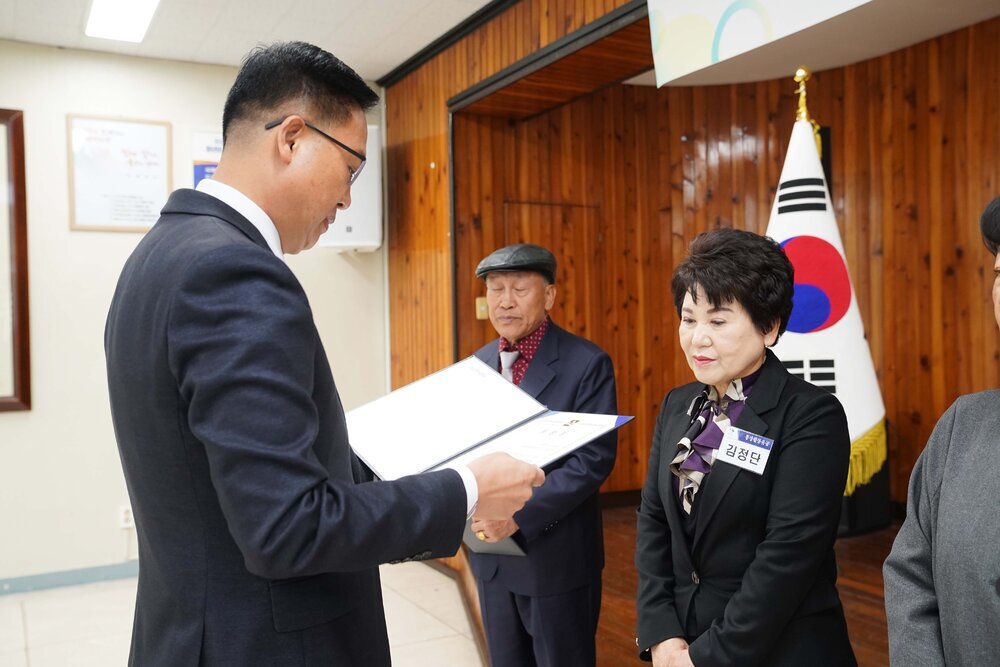 11월 정례조회로 정장을 입고 안경을 쓴 남성이 짧은 머리에 보라색블라우스를 입고 검은 정장을 입은 김정단 수상자에게 상장을 수여 하고 있다.