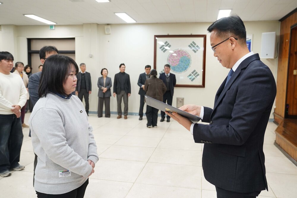 2026년 상반기 4급이하 정기인사로 정장을 입고 안경을 쓴 남성이 실내 행사장에서 한 여성에게 상장을 전달하며 내용을 읽고 있고, 여성은 회색 맨투맨을 입고 두 손을 모은 채 서 있으며 뒤쪽에는 여러 사람들이 줄지어 서서 행사를 지켜보고 있다.