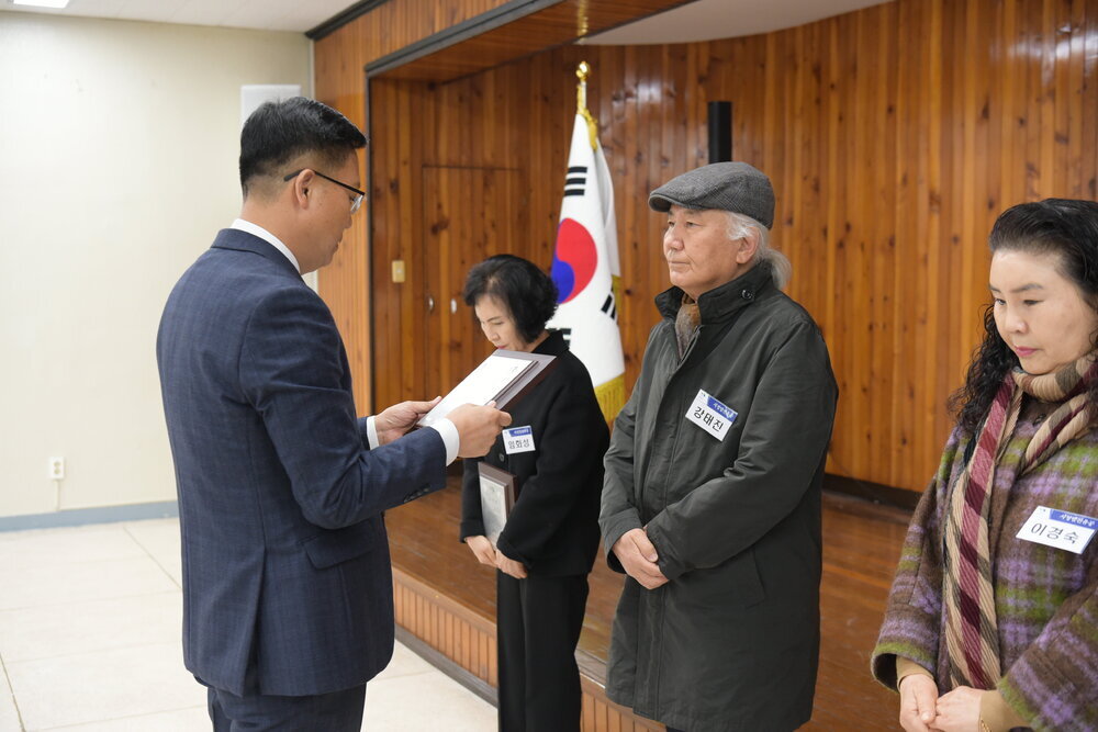 3월 정례조회에 정장을 입고 안경을 낀 남성이 흰머리 위로 회색 체크 모자를 쓰고 짙은 회색 반코트패딩을 입은 강태진에게 패를 수여하고 있다.