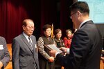 정장 차림의 관계자가 정장을 입은 남성 수상자에게 패를 전달하며 읽고 있는 모습이다.