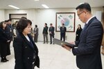 2026년 상반기 4급이하 정기인사로 정장을 입은 남성이 실내 행사장에서 한 여성에게 상장을 전달하며 내용을 읽고 있고, 여성은 짧은 파마머리에 검은색 트위드 자켓을 입고 두 손을 내린 상태로 서 있으며 뒤쪽으로 참석자들이 줄지어 서서 행사를 지켜보고 있다.