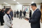 2026년 상반기 4급이하 정기인사로 정장을 입은 남성이 실내 행사장에서 한 여성에게 상장을 전달하며 내용을 읽고 있고, 여성은 검은색 목폴라 위에 밝은 회색 자켓을 입고 사원증을 착용한 채 두 손을 모은 상태로 서 있으며 뒤쪽으로 참석자들이 행사를 지켜보고 있다.