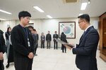 2026년 상반기 4급이하 정기인사로 정장을 입은 남성이 실내 행사장에서 한 남성에게 상장을 전달하며 내용을 읽고 있고, 남성은 검은 맨투맨을 입은 채 두 손을 모은 상태로 서 있으며 뒤쪽에는 참석자들이 줄지어 서서 행사를 지켜보고 있다.