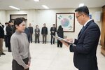 2026년 상반기 4급이하 정기인사로 정장을 입고 안경을 쓴 남성이 실내 행사장에서 한 여성에게 상장을 전달하며 내용을 읽고 있고, 여성은 회색 니트를 입고 두 손을 모은 채 서 있으며 뒤쪽에는 참석자들이 줄지어 서서 행사를 지켜보고 있다.